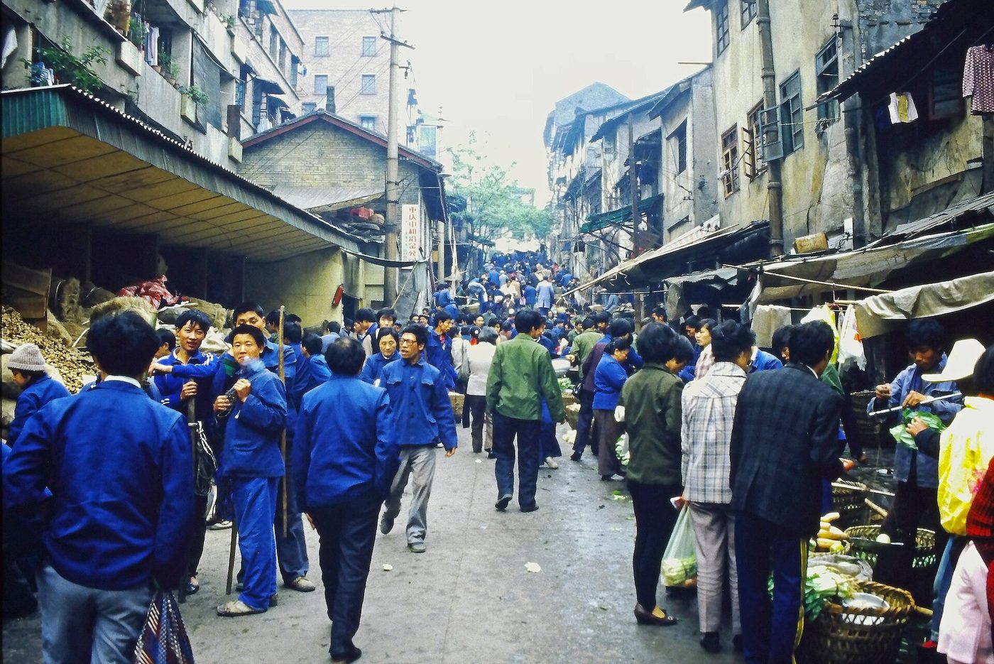 A busy street market in Chongqing in 1988, with low buildings, crowds and produce stalls.
